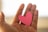 A hand is shown holding a small pink paper heart against a softly lit background, implying a gesture of giving or receiving love.
