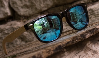 A pair of sunglasses with black frames and wooden arms is placed on a wooden surface. The reflective lenses capture a scene of an ancient stone wall pathway with lush greenery, presenting a blend of natural and architectural elements.