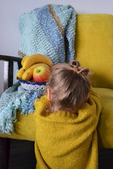 A young child with two small buns in their hair is leaning against a yellow armchair. A bowl of fruit, including bananas and apples, rests on the armchair. There is a textured blue and brown blanket draped over the chair.
