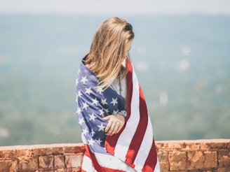 woman standing leaning on brick wall wrapping body with U.S. flag near body of water onlyfans