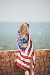 woman standing leaning on brick wall wrapping body with U.S. flag near body of water onlyfans