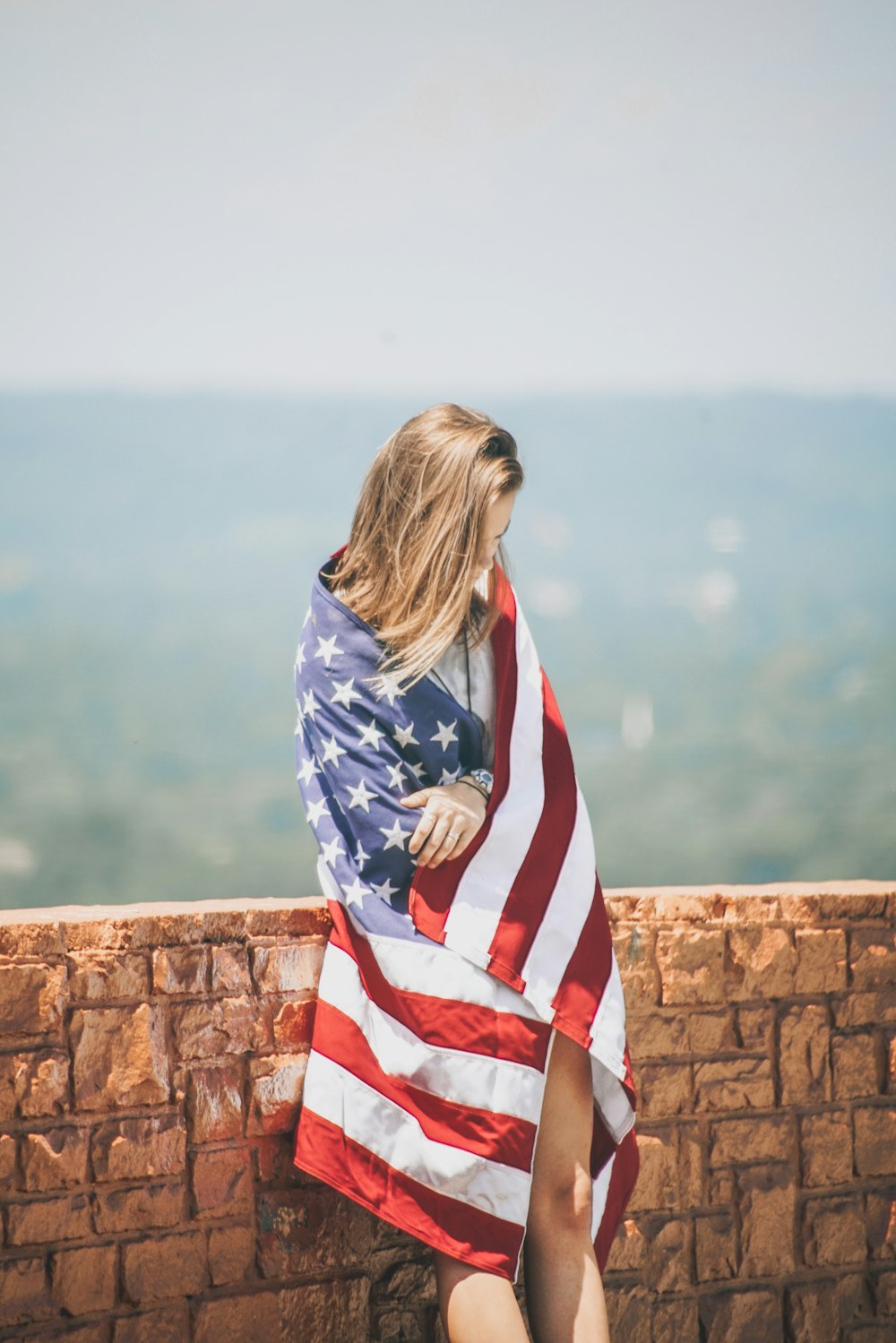 mujer de pie apoyada en la pared de ladrillo que envuelve el cuerpo con la bandera de EE. UU. cerca del cuerpo de agua