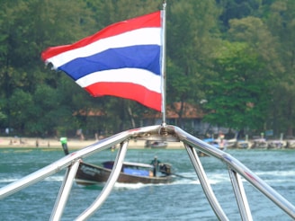 A red, white, and blue flag waves prominently on a boat, with a lush, green forested shoreline and a beach in the background. A smaller boat passes by on the water, leaving a trail behind.
