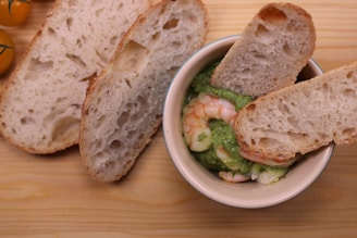 A bowl filled with shrimp and a green sauce, likely guacamole, garnished with slices of crusty bread. The bowl is placed on a wooden surface alongside a few yellow cherry tomatoes on the vine.