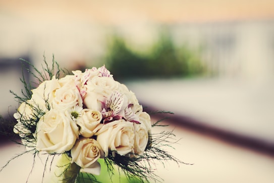 A close-up bouquet of cream-colored roses with delicate detailing of pink and white lilies mixed in, surrounded by soft green foliage. The background is blurred, creating a dreamy, out-of-focus effect, emphasizing the elegance of the flowers.