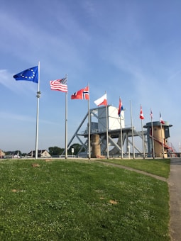 A row of flagpoles displaying various national flags stands on a grassy hill. In the background, there is a large industrial structure resembling a bridge or dam, and a red and white tower with a spiral staircase.