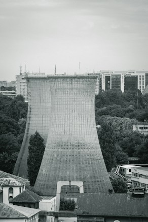 A monochromatic scene featuring two large cooling towers in an urban landscape, surrounded by a mix of industrial and office buildings. Trees and foliage are visible at the base, adding a touch of nature to the otherwise industrial setting. The sky is overcast, suggesting a moody atmosphere.