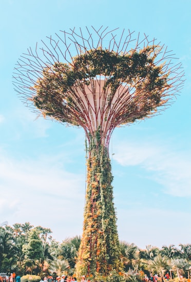 A towering structure resembling a futuristic tree, adorned with lush greenery and foliage climbing up its trunk. The top is capped with a canopy-like design, featuring a network of radial branches extending outwards. The base of the structure is surrounded by tropical plants and a gathering of people, suggesting a public or tourist area. The sky above is clear and blue, contributing to a vibrant and lively atmosphere.