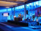A spacious room with a modern interior design features a stage where a person is giving a presentation. Large windows line the walls, allowing blue light to fill the space. Several large screens around the room display graphics. There are potted plants and chairs adding to the decor.