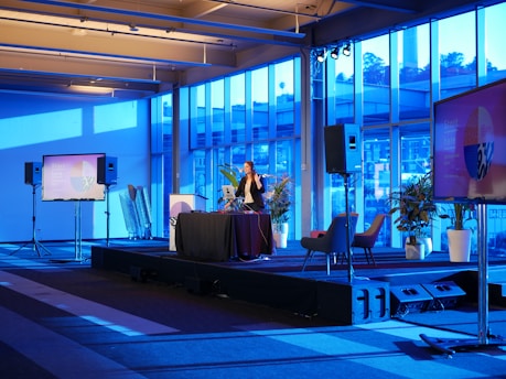 A spacious room with a modern interior design features a stage where a person is giving a presentation. Large windows line the walls, allowing blue light to fill the space. Several large screens around the room display graphics. There are potted plants and chairs adding to the decor.