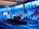 A spacious room with a modern interior design features a stage where a person is giving a presentation. Large windows line the walls, allowing blue light to fill the space. Several large screens around the room display graphics. There are potted plants and chairs adding to the decor.