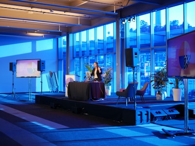 A spacious room with a modern interior design features a stage where a person is giving a presentation. Large windows line the walls, allowing blue light to fill the space. Several large screens around the room display graphics. There are potted plants and chairs adding to the decor.