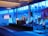 A spacious room with a modern interior design features a stage where a person is giving a presentation. Large windows line the walls, allowing blue light to fill the space. Several large screens around the room display graphics. There are potted plants and chairs adding to the decor.