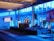A spacious room with a modern interior design features a stage where a person is giving a presentation. Large windows line the walls, allowing blue light to fill the space. Several large screens around the room display graphics. There are potted plants and chairs adding to the decor.