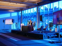 A spacious room with a modern interior design features a stage where a person is giving a presentation. Large windows line the walls, allowing blue light to fill the space. Several large screens around the room display graphics. There are potted plants and chairs adding to the decor.