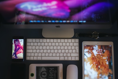 A neatly organized workspace with a focus on electronic devices. At the top, a desktop computer monitor displays a blurred image, possibly reflecting colorful lights. The foreground is dominated by a white keyboard and mouse, flanked by a smartphone on the left showing a lock screen with a time of 20:11, and a tablet on the right with a background featuring autumn leaves, showing the time as 20:17. A watch lies above the tablet, and there is an e-reader partially visible below the phone and keyboard.