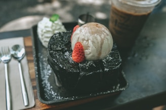 A delectable dessert featuring a scoop of vanilla ice cream drizzled with chocolate syrup, placed atop a thick slice of dark chocolate cake garnished with fresh strawberries. Beside it, a dollop of whipped cream with a mint leaf, and in the background, a glass of iced coffee. The scene is elegantly arranged on a tray with polished silverware.