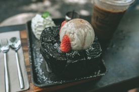 A delectable dessert featuring a scoop of vanilla ice cream drizzled with chocolate syrup, placed atop a thick slice of dark chocolate cake garnished with fresh strawberries. Beside it, a dollop of whipped cream with a mint leaf, and in the background, a glass of iced coffee. The scene is elegantly arranged on a tray with polished silverware.