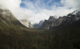 A scenic valley surrounded by towering mountains with snow-capped peaks. A tall waterfall cascades down the mountain on the right side, with dense forests covering the valley floor. Clouds partially cover the sky, adding a mystical atmosphere to the landscape.