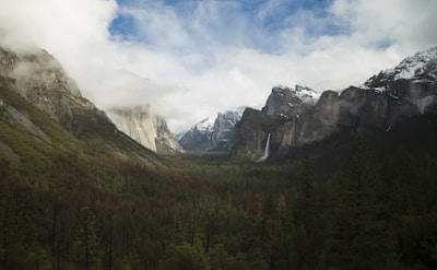 A scenic valley surrounded by towering mountains with snow-capped peaks. A tall waterfall cascades down the mountain on the right side, with dense forests covering the valley floor. Clouds partially cover the sky, adding a mystical atmosphere to the landscape.
