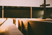 A dimly lit interior of a church with wooden pews illuminated by warm sunlight. The light creates a serene atmosphere and a large wooden cross is visible at the front.