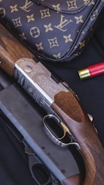 A luxury handbag with a monogram pattern is displayed alongside a detailed shotgun with engraved metal and wooden components. There is a red shotgun shell nearby, set against a dark, textured background.