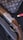 A luxury handbag with a monogram pattern is displayed alongside a detailed shotgun with engraved metal and wooden components. There is a red shotgun shell nearby, set against a dark, textured background.