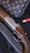 A luxury handbag with a monogram pattern is displayed alongside a detailed shotgun with engraved metal and wooden components. There is a red shotgun shell nearby, set against a dark, textured background.