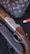 A luxury handbag with a monogram pattern is displayed alongside a detailed shotgun with engraved metal and wooden components. There is a red shotgun shell nearby, set against a dark, textured background.