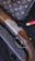 A luxury handbag with a monogram pattern is displayed alongside a detailed shotgun with engraved metal and wooden components. There is a red shotgun shell nearby, set against a dark, textured background.