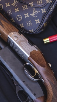 A luxury handbag with a monogram pattern is displayed alongside a detailed shotgun with engraved metal and wooden components. There is a red shotgun shell nearby, set against a dark, textured background.
