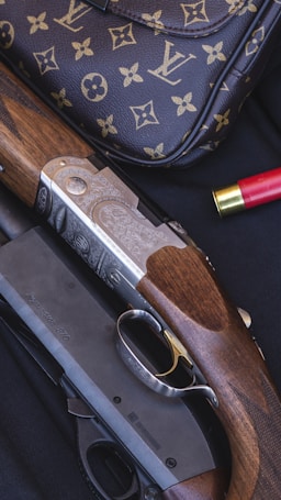 A luxury handbag with a monogram pattern is displayed alongside a detailed shotgun with engraved metal and wooden components. There is a red shotgun shell nearby, set against a dark, textured background.