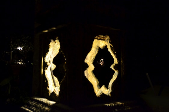 An artistic lamp features intricate cut-out patterns illuminated from within, casting a warm glow. The background is dark, suggesting an evening or nighttime setting. The light creates interesting silhouettes and shadows, enhancing the detailed craftsmanship of the lamp.