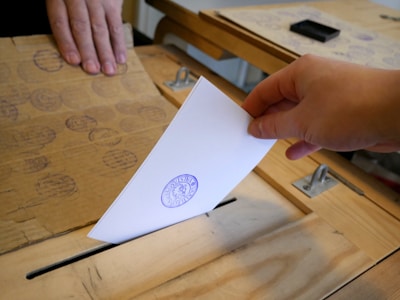 A hand is inserting a ballot or document into a wooden box that resembles a ballot box. Another hand is holding the box steady. There is a visible stamp on the paper being inserted.