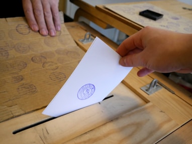 A close-up of a hand placing a ballot into a transparent voting box.