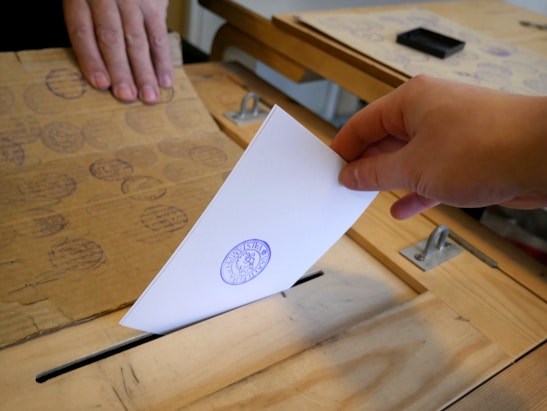 Close-up of a hand casting a vote into a ballot box with a blurred crowd in the background.