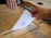 A hand is inserting a ballot or document into a wooden box that resembles a ballot box. Another hand is holding the box steady. There is a visible stamp on the paper being inserted.