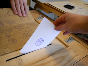 A close-up of hands dropping a ballot into a transparent voting box.