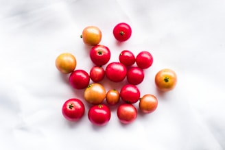A collection of small, glossy red and orange fruits are scattered across a plain white surface. The fruits appear fresh, with varying shades of ripeness, from bright red to light orange, and small green stems are visible on some.