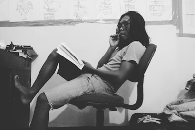 A person with long hair and glasses is seated on a chair, holding a book and contemplating. The setting appears to be a study room with flowcharts or diagrams on the wall in the background. The person is dressed casually in a t-shirt and shorts, and there are various papers or books on a desk nearby.