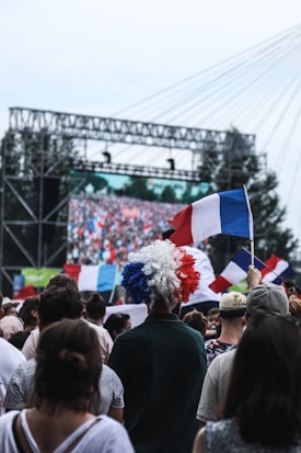 A large crowd of people gathered outdoors, many holding French flags and wearing patriotic attire. A person with a tricolor wig stands out in the foreground. In the background, a large screen displays an image of a packed stadium or crowd scene, indicating a public event or celebration. Trees and a structure resembling a bridge or stage rigging are visible.