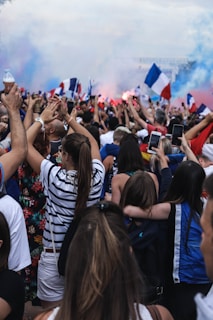A large crowd of people is gathered outdoors in a celebratory atmosphere. Many individuals are holding up smartphones, taking photos or videos, and waving flags. The flags prominently displayed are French flags, with blue, white, and red colors. Smoke or fog, tinged with colored light, fills the background, adding to the festive environment. The crowd appears densely packed, including a mix of men and women with excited expressions.