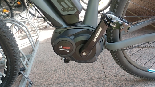 A close-up view of a bicycle's pedal and crank area, featuring a Bosch Performance Line CX motor. The bike is parked in a metallic bike rack. The surrounding surface is a tiled pavement with other bicycles partially visible in the background.