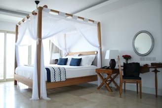 A well-lit and modern bedroom features a four-poster wooden bed with white drapes and navy blue pillows. The room includes a wooden desk with a matching chair, adorned with a lamp and a vase of red roses. A large round mirror hangs on the wall above the desk. The flooring appears to be marble, and there is natural light coming through the large windows.