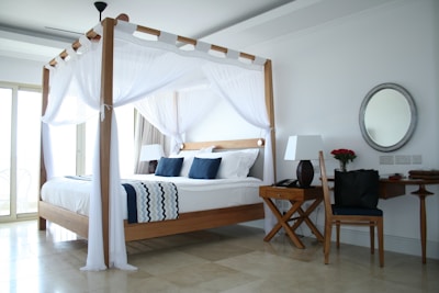 A well-lit and modern bedroom features a four-poster wooden bed with white drapes and navy blue pillows. The room includes a wooden desk with a matching chair, adorned with a lamp and a vase of red roses. A large round mirror hangs on the wall above the desk. The flooring appears to be marble, and there is natural light coming through the large windows.