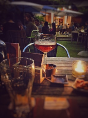 A cozy outdoor setting at a restaurant or bar, with a focus on a cocktail glass half-filled with amber-colored liquid. The background features people sitting at tables and enjoying their evening under large umbrellas, while a lit candle adds to the intimate ambiance. There are snacks on the table, contributing to the relaxed atmosphere.
