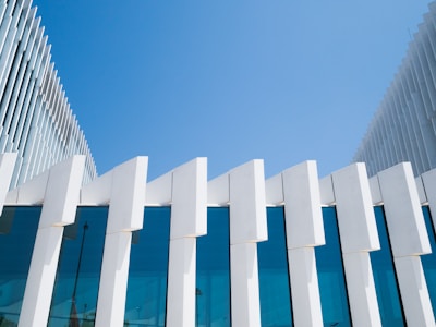 A modern architectural structure featuring a facade with large, vertical white concrete panels and clear blue-tinted glass windows. The building design includes sharp geometric lines and angles, with a clear blue sky in the background, adding to its contemporary aesthetic.