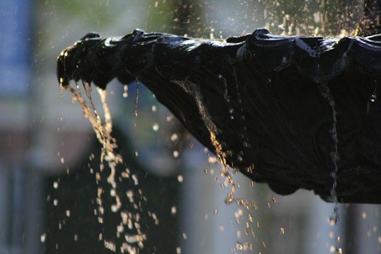 A dark, intricately designed fountain spills water gently over its edge, with sunlight catching the droplets, creating a shimmering effect. The background is softly blurred, emphasizing the focus on the water flow.