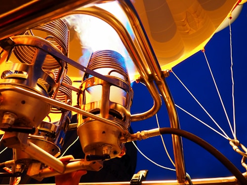The image features a close-up view of a hot air balloon burner mechanism. Metal components and flames are prominent, with a background showcasing the underside of the balloon's fabric against a deep blue sky.