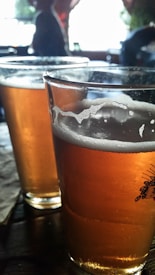 Two glasses filled with amber-colored beer sit on a dark wooden table. The glasses have a thin layer of frothy foam along the top. The background appears to be a softly blurred bar or pub setting with some indistinct figures.
